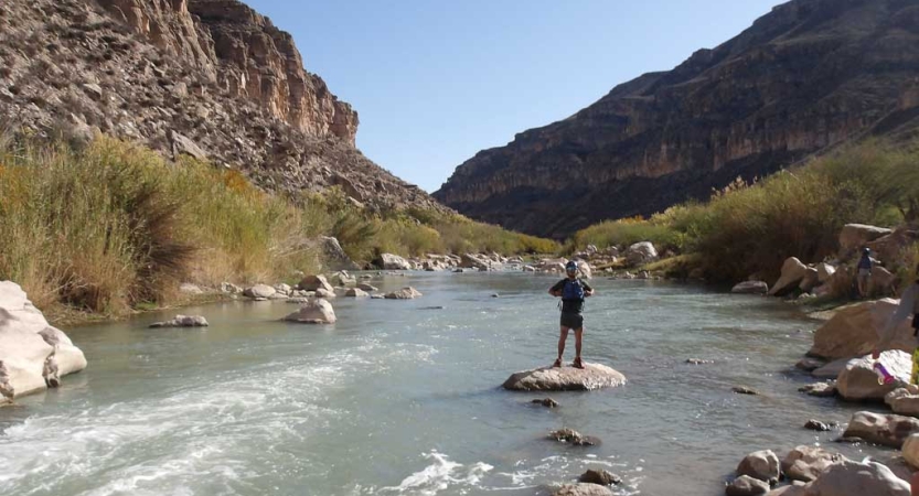 A person wearing a life jackets stands on rock in the middle of a river. The river is framed by high canyon walls. 